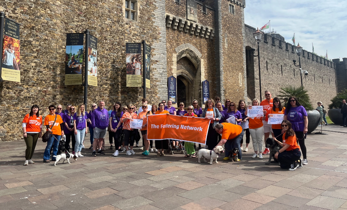 A group of people in orange and purple t-shirts stand together holding an orange 'The Fostering Network' banner. 
